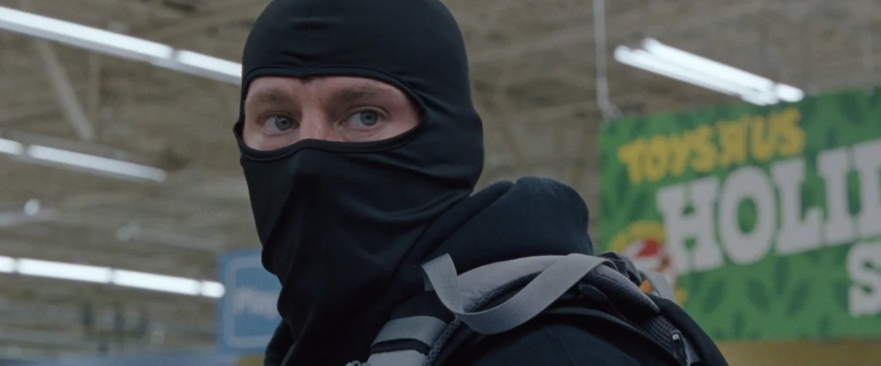 Roofman (2025) movie still: closeup — A man wearing a black balaclava stands inside a brightly lit retail store aisle. He wears a dark hoodie and…