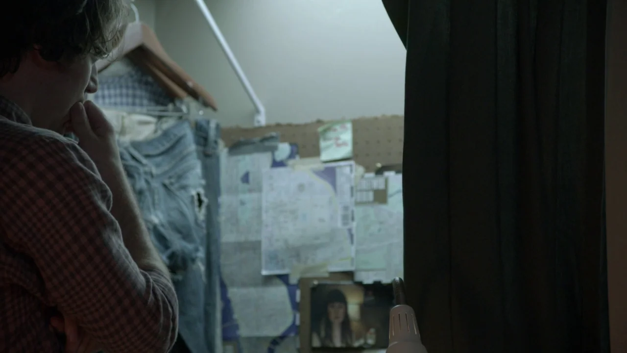 The Heart Machine (2014) movie still: medium — A small, dimly lit closet features a wall covered in maps and taped notes. A man stands in the…