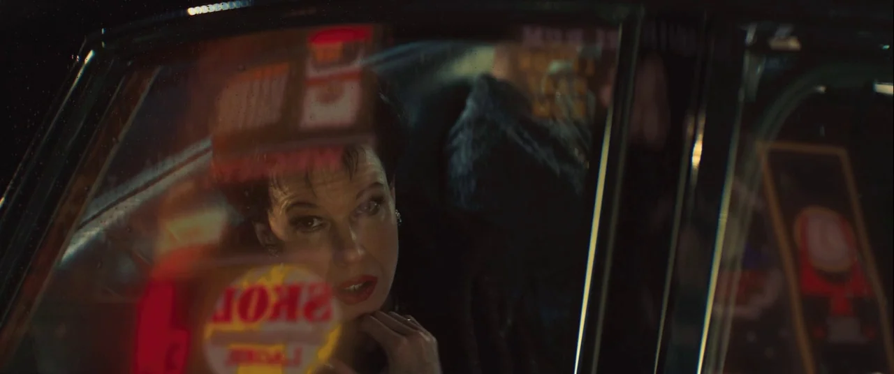 Judy (2019) movie still: closeup — A woman looks through the window of a car at night. Red neon signage reflects against the glass pane obscuring…