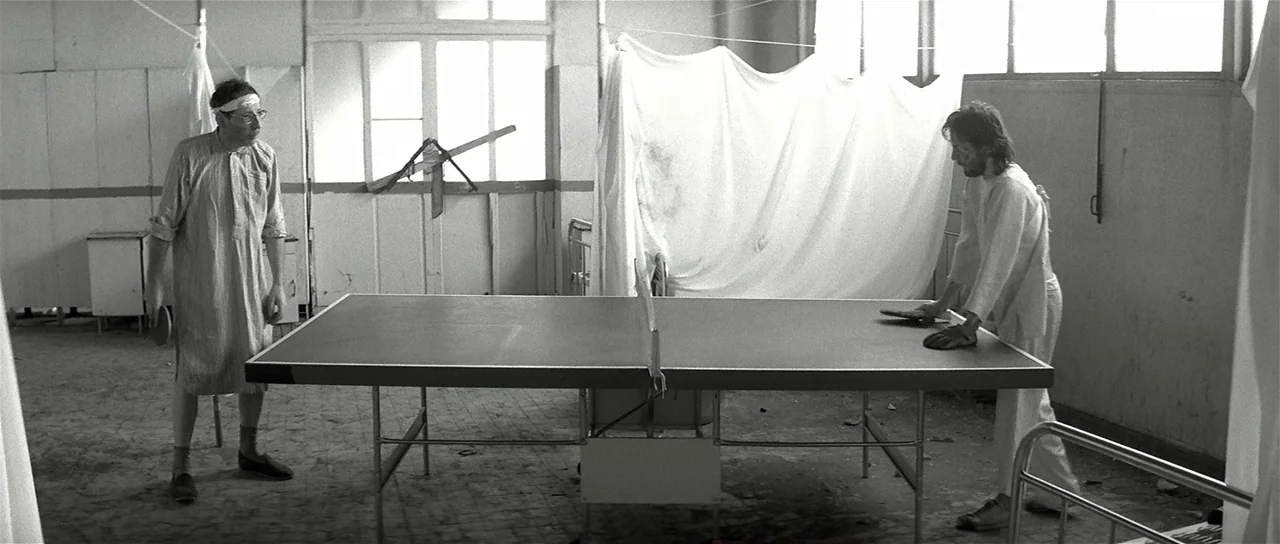 The Last Battle (1983) movie still: wide — A dilapidated room features peeling walls and makeshift white privacy curtains on frames. Two men in…