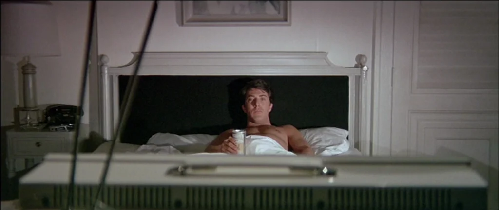 The Graduate (1967) movie still: medium — A dimly lit hotel room features a bed against a white headboard. A man lies in the bed holding a beer…