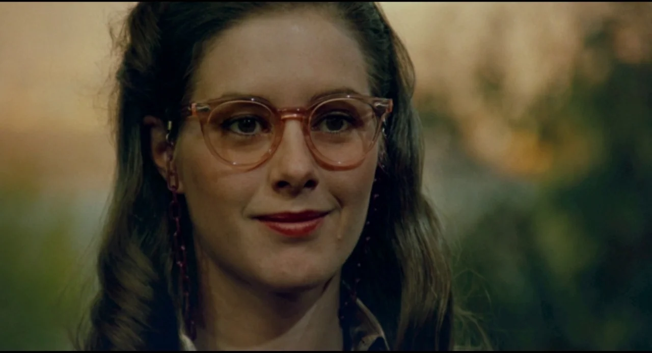 Olivia (1983) movie still: closeup — An outdoor setting features soft focus green foliage in the blurred background. A young woman with brown hair…