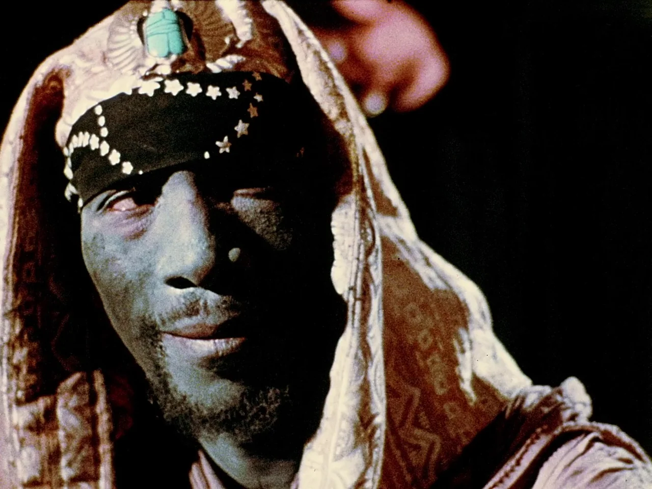 Space is the Place (1974) movie still: closeup — A close portrait of a man wearing a decorative headscarf with a scarab. He looks toward the side…