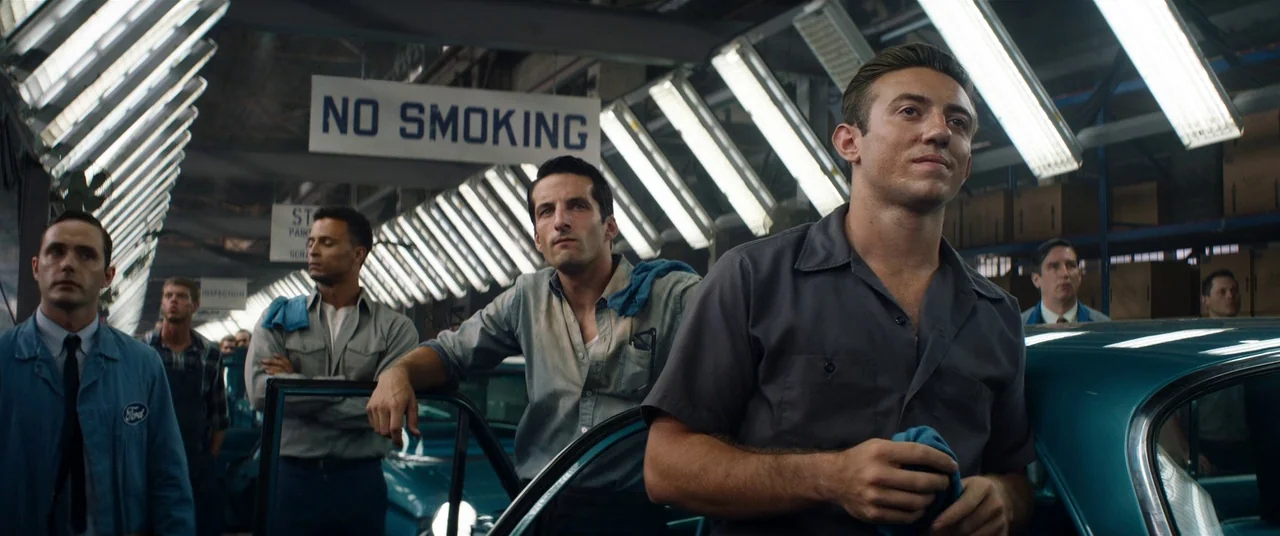 Ford V Ferrari (2019) movie still: medium — A bustling factory floor featuring a line of unfinished teal cars under overhead lights. Several men in…