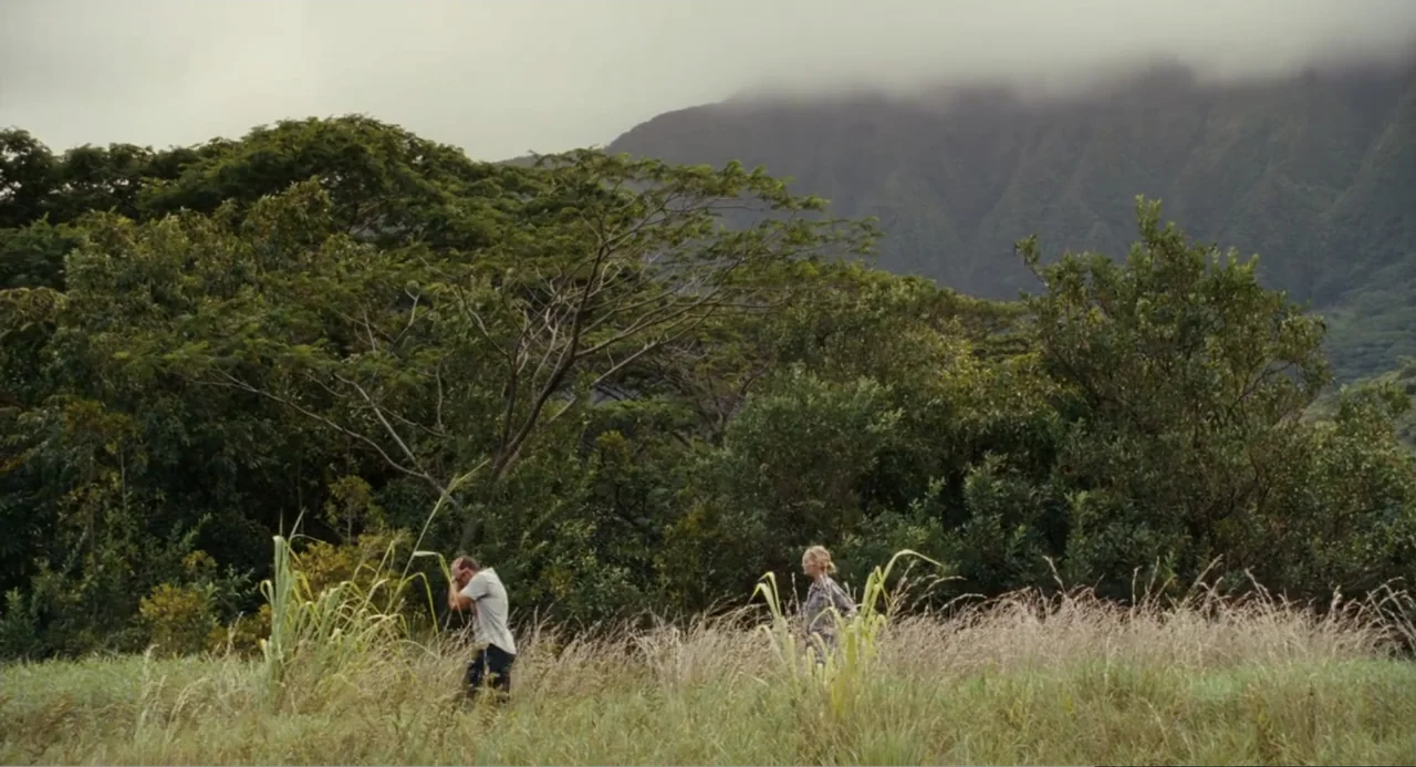 Aloha (2015) movie still: wide — A man and woman traverse an open grassy field beneath a clouded mountain range. The subjects move together toward a…