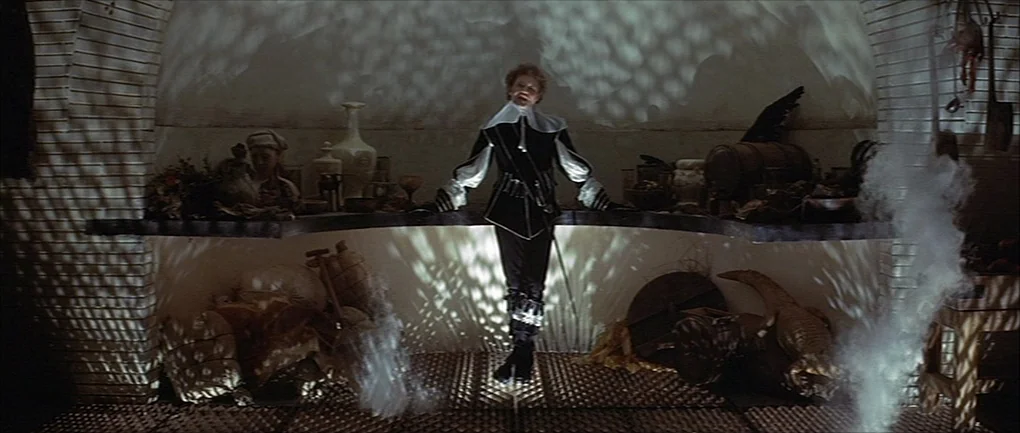 The Devils (1971) movie still: fullbody — A dark subterranean space features stone arches and clutter scattered across the floor. A man in period…