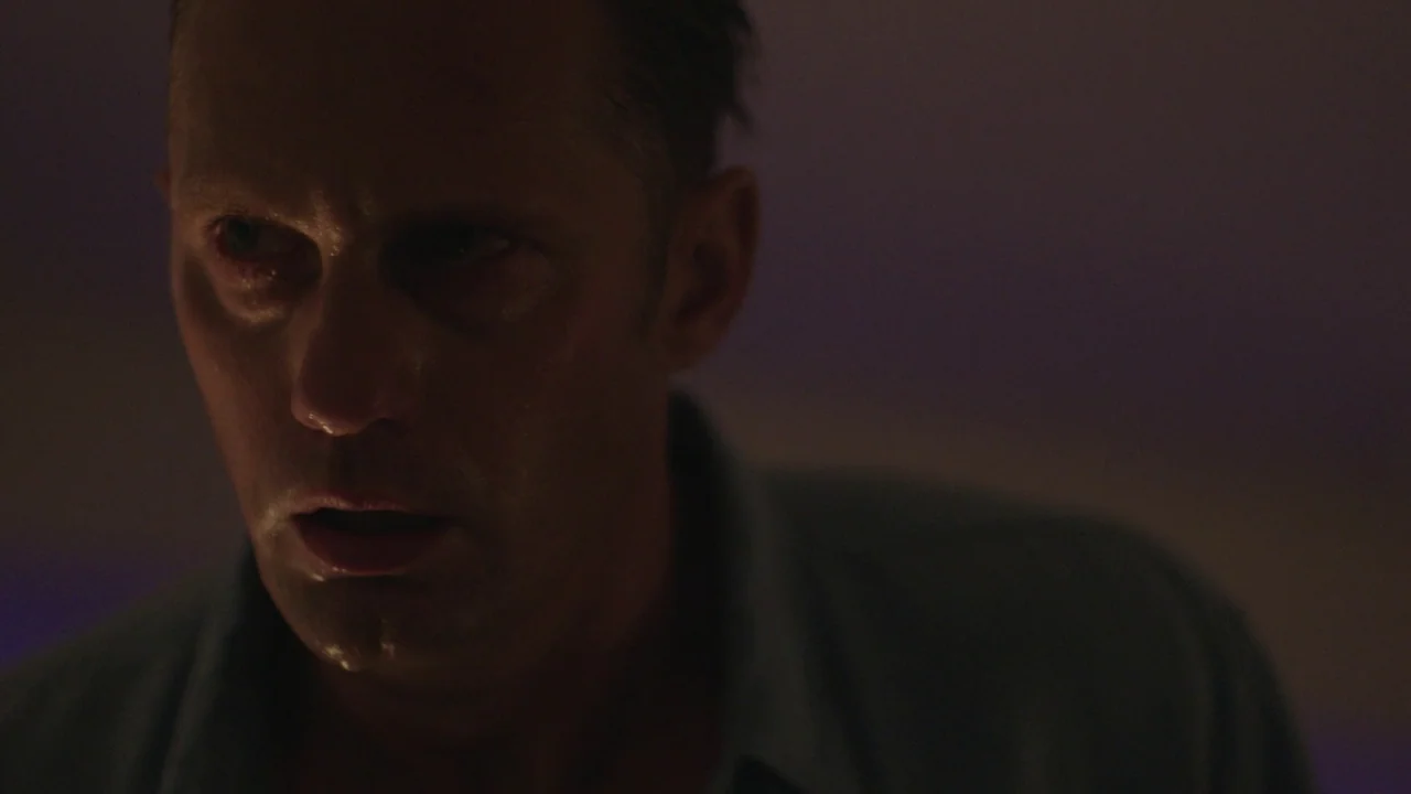 Infinity Pool (2023) movie still: closeup — A dimly lit room with heavy shadows obscures the background environment. A man with a damp face stares…