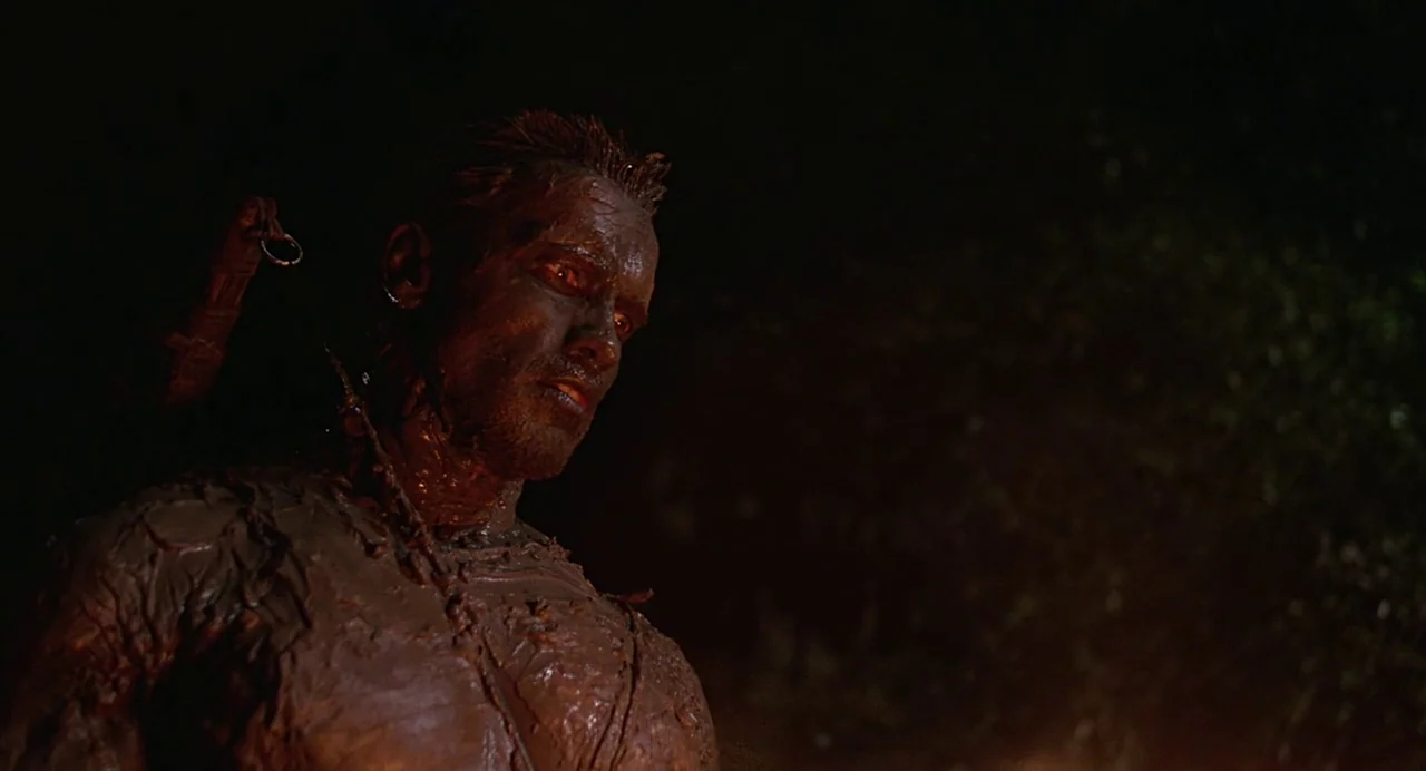 Predator (1987) movie still: closeup — A dark, dense jungle environment at night. A muscular man covered in mud looks down toward the ground.