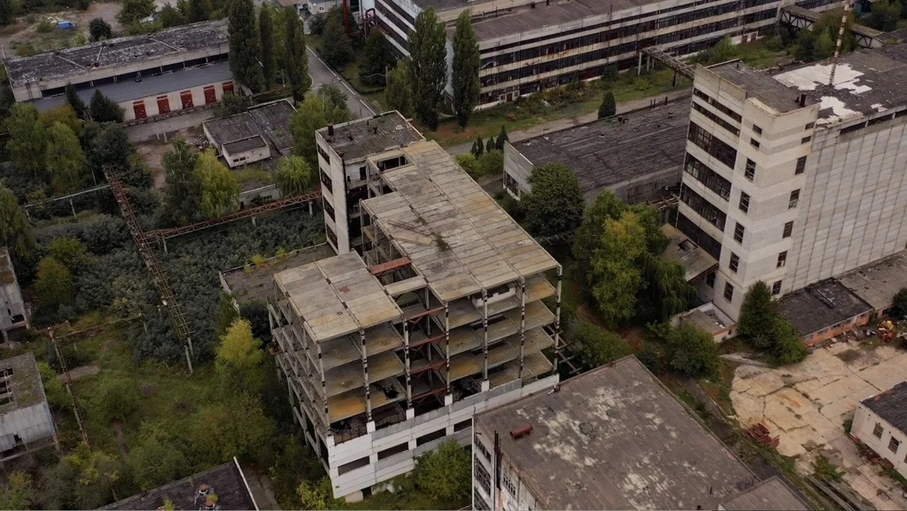 The Collective (2023) movie still: establishing — An abandoned industrial site features several multi-story concrete structures surrounded by…