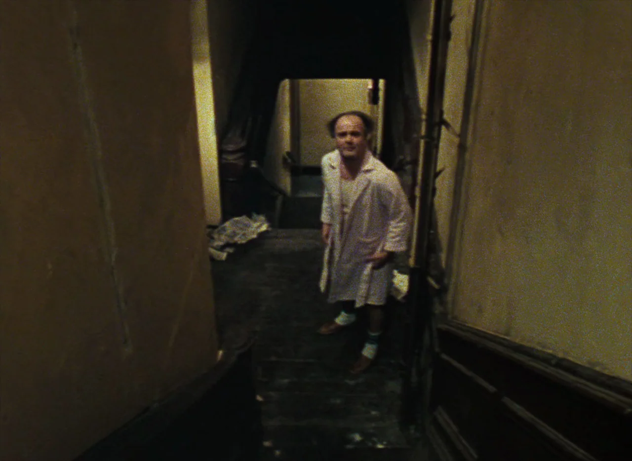 Basket Case (1982) movie still: fullbody — A dimly lit narrow indoor stairwell with dark wooden floorboards and beige walls. An adult male wearing a…