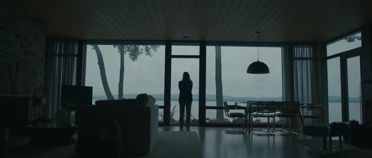 Arrival (2016) movie still: wide — A modern, minimalist house features large glass windows overlooking a vast, overcast lake. A woman stands in the…
