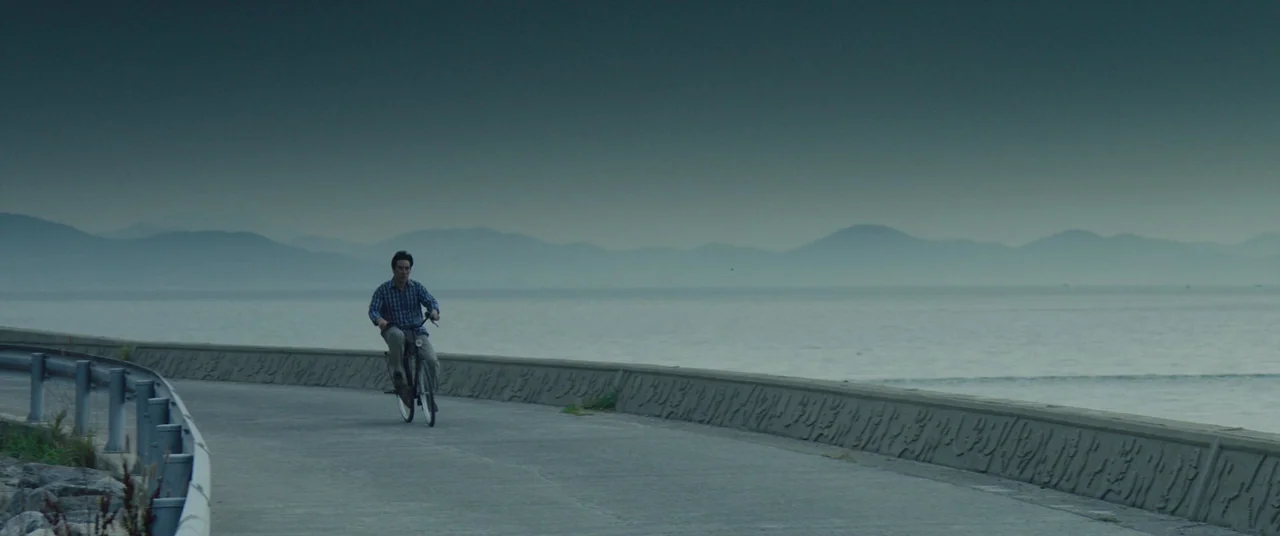 Beasts Clawing At Straws (2020) movie still: wide — A paved path runs along a concrete sea wall beside a calm, gray ocean. A man rides a bicycle…