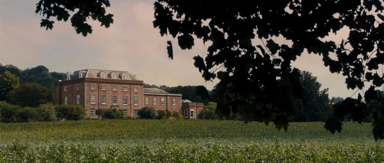 Road Games (2015) movie still: establishing — A multi-story brick manor house stands prominently behind a large, expansive green crop field.…