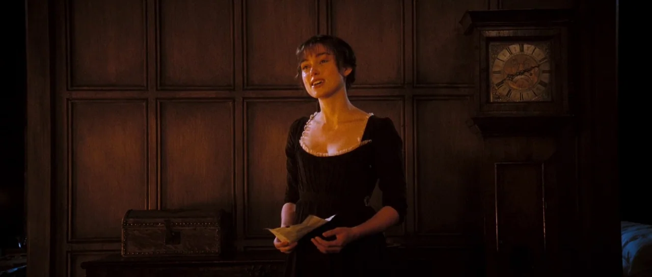 Pride & Prejudice (2005) movie still: medium — A dimly lit room features dark wood paneling and a large vintage standing clock. A young woman in a…
