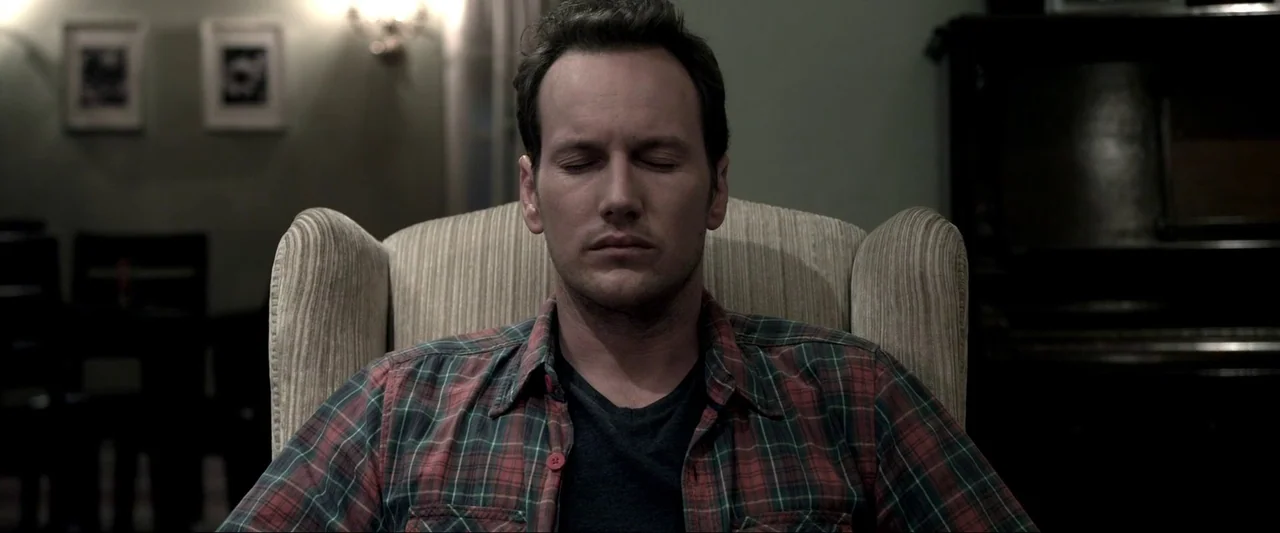 Insidious (2010) movie still: medium — A dimly lit room features a man sitting in a light-colored upholstered armchair. Two small framed pictures…