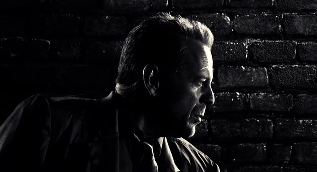 Sin City (2005) movie still: closeup — A man stands before a dark, textured brick wall in low light. He looks to the right side of the frame.
