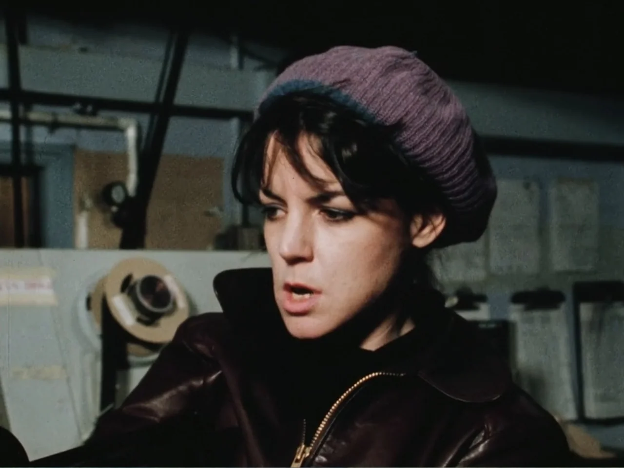 Born in Flames (1983) movie still: closeup — An interior production space with equipment visible in the blurred background. A woman in a leather…