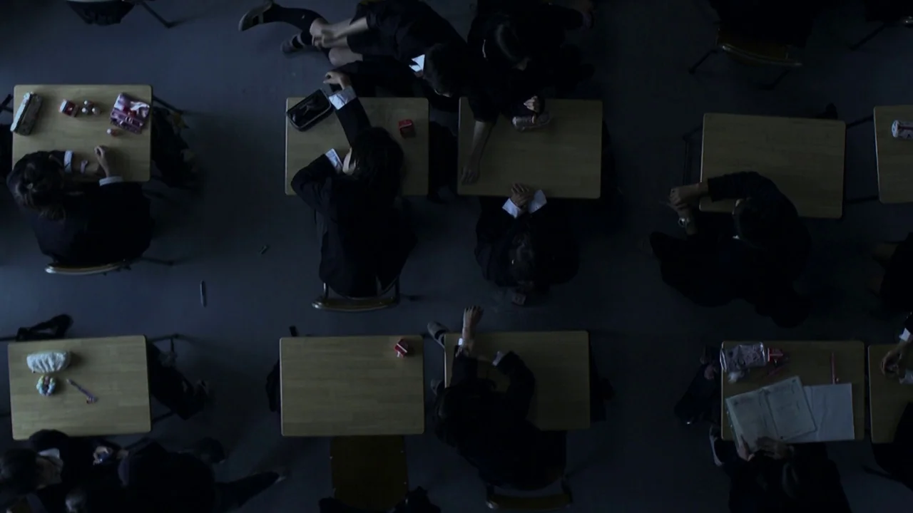 Confessions (2010) movie still: wide — A dimly lit classroom features rows of wooden desks arranged in a grid pattern. Several students in dark…