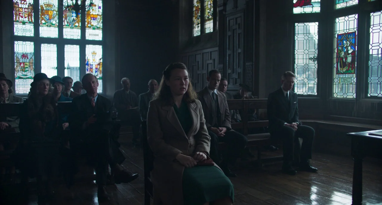 The Little Stranger (2018) movie still: medium — A group of people sit on wooden benches inside a dimly lit stone church. A woman in a tan coat sits…