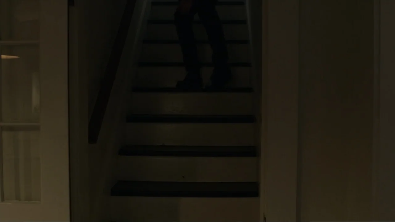The Collective (2023) movie still: medium — A dark, dimly lit indoor staircase features wooden steps and a white door frame. A person walks down the…