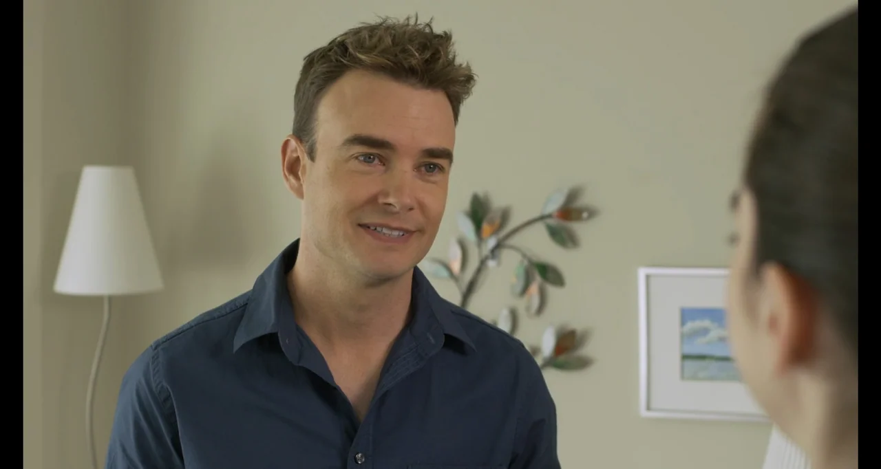 A.R.C.H.I.E. (2016) movie still: medium — A light-walled interior features a lamp and wall art in the background. A man in a blue shirt turns toward…