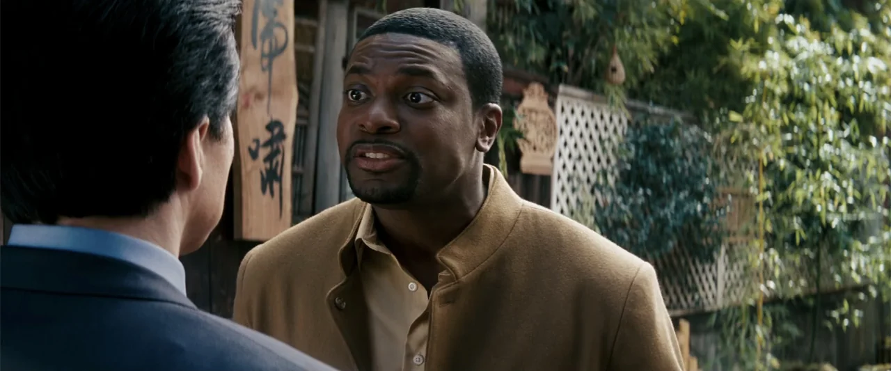 Rush Hour 3 (2007) movie still: medium — A man stands outdoors talking to another man in a suit. Green foliage is visible on the wall behind them.