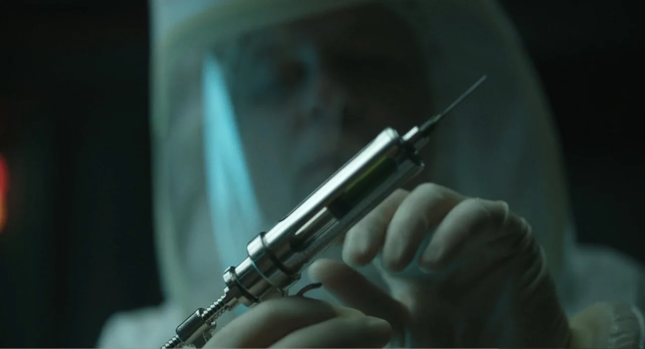 Mickey 17 (2025) movie still: closeup — A dimly lit sterile laboratory interior features a person in a protective white hazmat suit. The individual…
