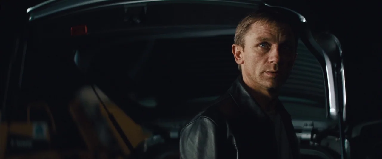 Layer Cake (2004) movie still: medium — A man stands before the open hatch of a dark car at night. He is positioned to the right side of the frame…