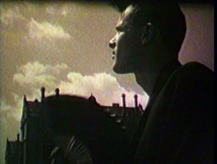 The Angelic Conversation (1985) movie still: medium — A man stands on a rooftop against a cloudy, light-filled sky. Behind him, the roofline of a…