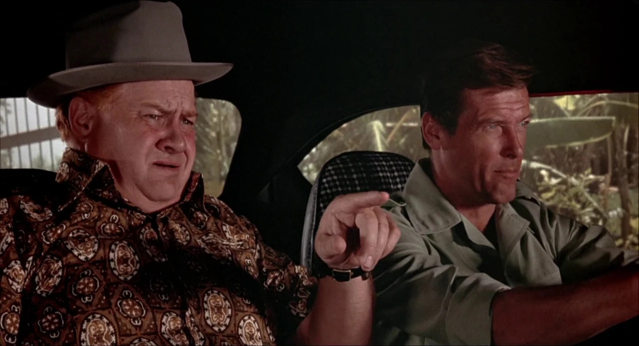 The Man With the Golden Gun (1974) movie still: medium — Inside a moving car, two men are seated side-by-side in the front seats. The passenger…