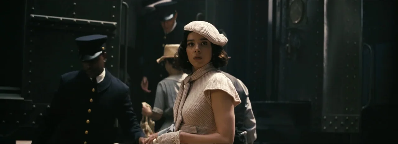Sinners (2025) movie still: medium — A 1920s train station platform features dark metal train cars and blurred passengers. A young woman in a beige…