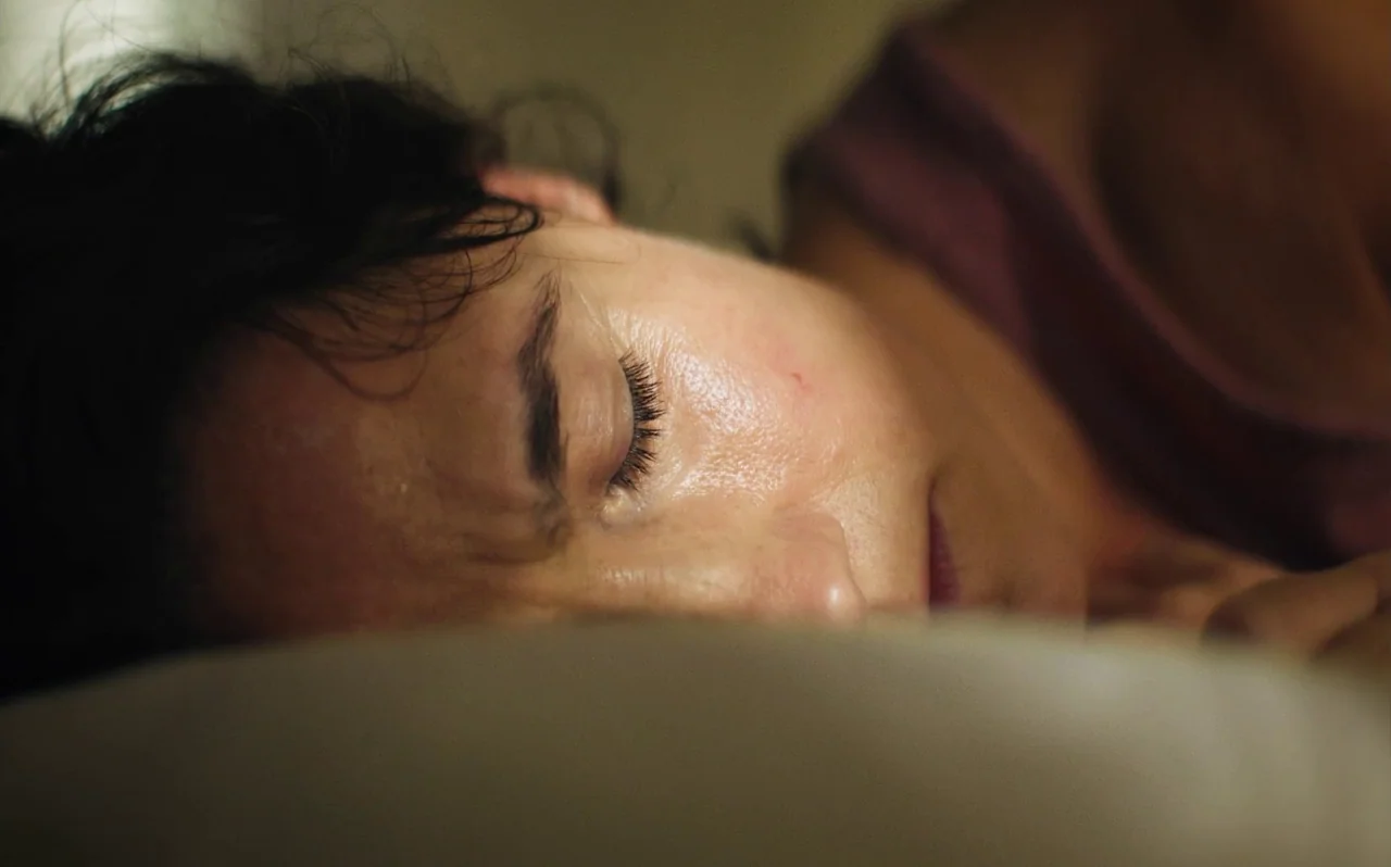 Knocking (2021) movie still: closeup — A person rests their head on a soft, light-colored pillow in darkness. The subject lies with eyes closed…