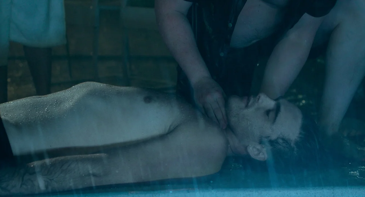 Deep Water (2022) movie still: A somber, tense moment captured in the dark of night where a man lies prone in shallow water, surrounded by rain. The…