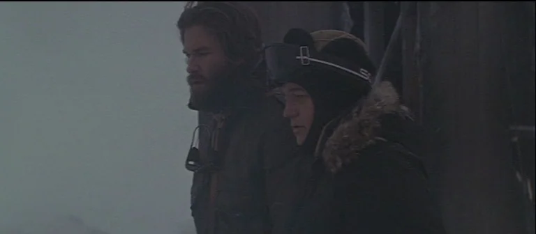 The Thing (1982) movie still: medium — An exterior snowy landscape obscured by heavy overcast conditions and fog. Two men in winter gear stand close…