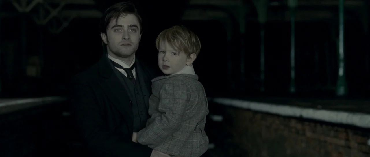 The Woman in Black (2012) movie still: medium — A man and child stand on a dark, desolate train platform at night. The man carries the young boy as…