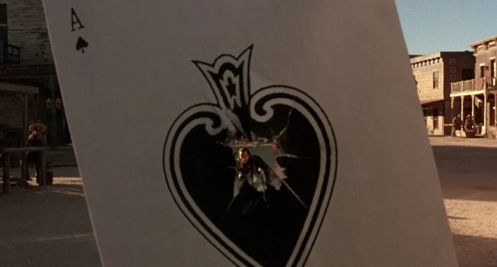 The Quick and the Dead (1995) movie still: closeup — An ace of spades playing card with a large bullet hole held in the foreground. Through the hole…