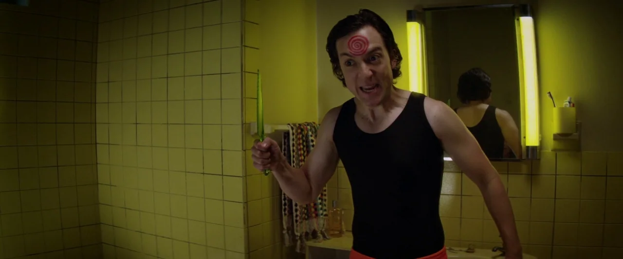 Seven Stages to Achieve Eternal Bliss (2018) movie still: medium — A tiled bathroom features yellow walls and vertical fluorescent light strips…