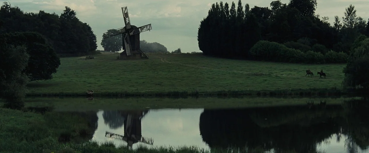 War Horse (2011) movie still: establishing — A rolling green field features an old wooden windmill standing near a calm pond. Two riders on…