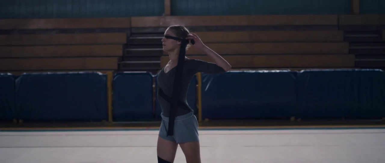 Alps (2011) movie still: medium — A spacious indoor sports facility features wooden bleachers in the background and a mat. A young woman in athletic…