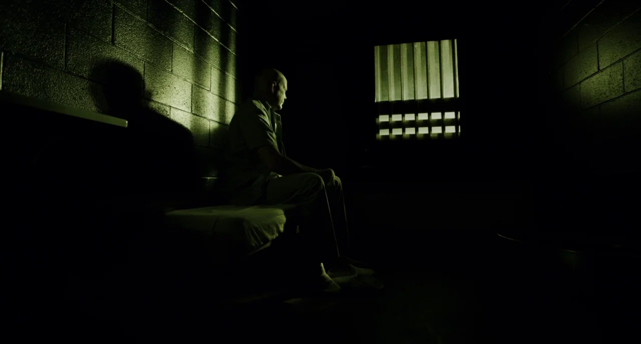 Brawl in Cell Block 99 (2017) movie still: fullbody — A dark, spartan room features cinder block walls and a thin mattress on a cot. A bald man sits…