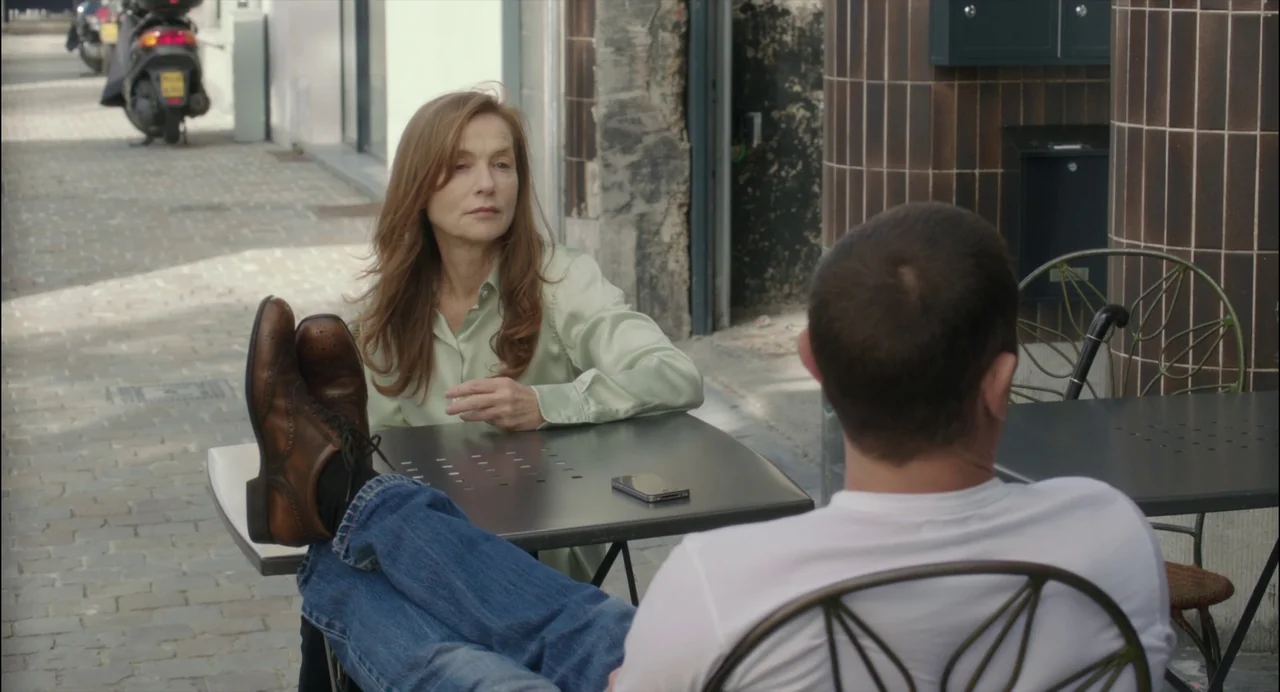 Abuse of Weakness (2013) movie still: medium — A sidewalk cafe area with paved stone ground and a metal-tiled building facade. A woman sits across…