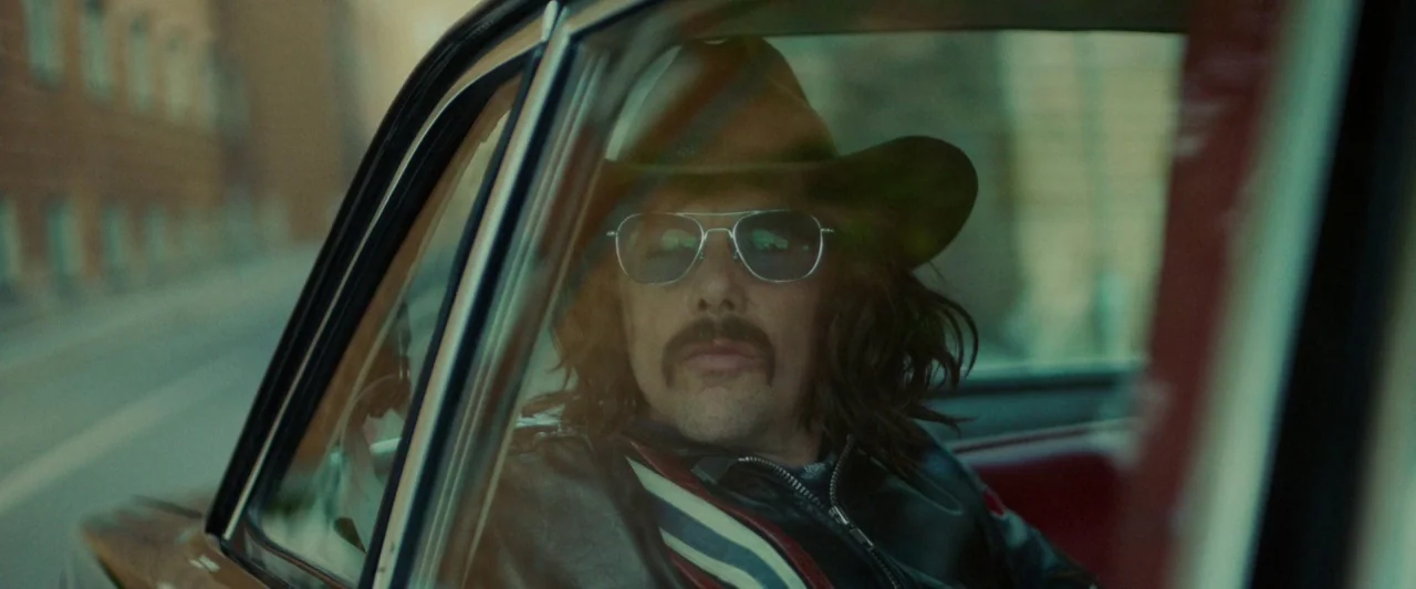 Stockholm (2019) movie still: closeup — A man sits inside a vehicle while looking through the side window. He wears a cowboy hat and a leather…