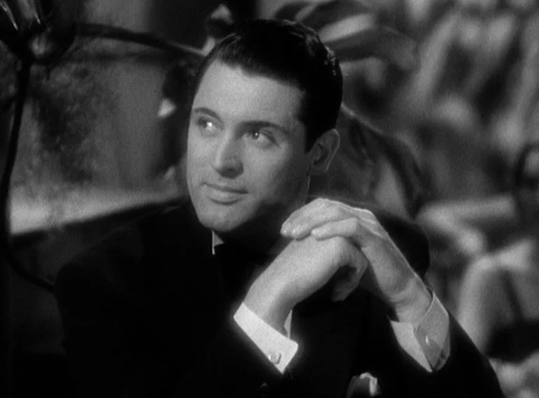 Blonde Venus (1932) movie still: closeup — A man in a tuxedo sits at a table in a dimly lit setting. He keeps his hands clasped as he looks…