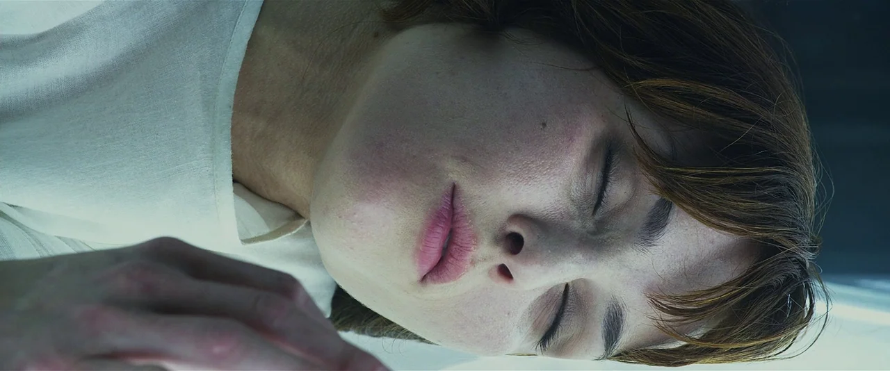 Prometheus (2012) movie still: closeup — A close-up shot focuses on the face of a woman lying down. The background is indistinct and dark.