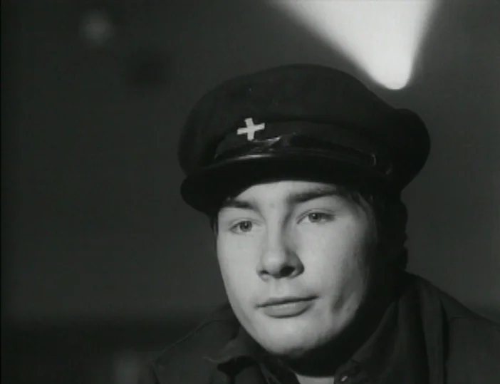 Les Carabiniers (1963) movie still: closeup — A dimly lit studio environment with a dark background and a single light streak. The man wears a dark…