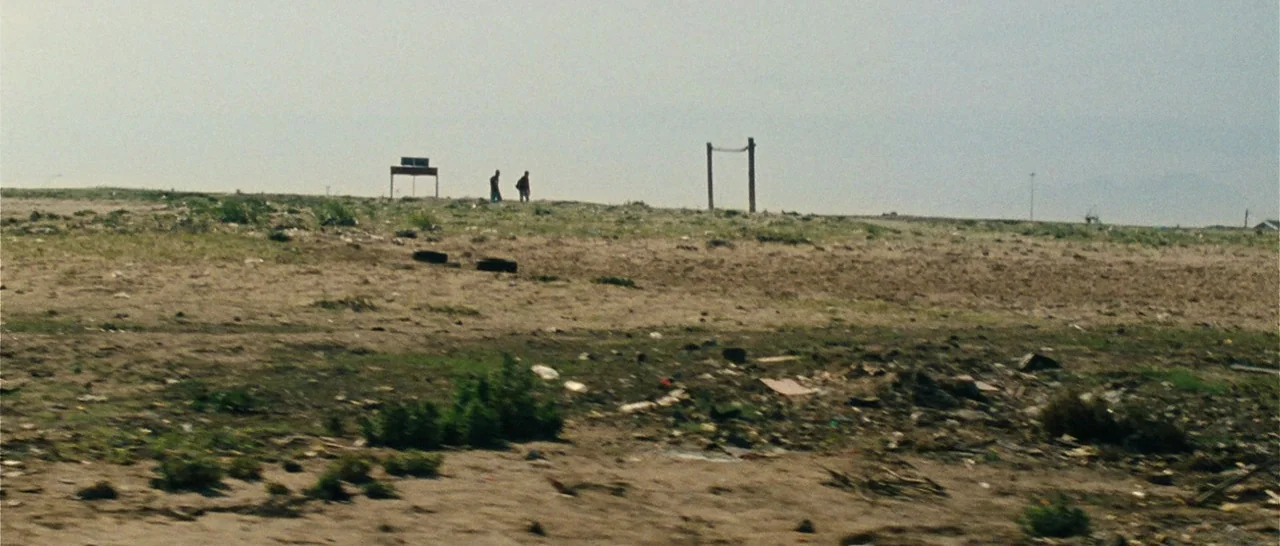 Message From The King (2016) movie still: wide — A desolate expanse of dirt and sparse weeds contains scattered debris and litter. Two distant…