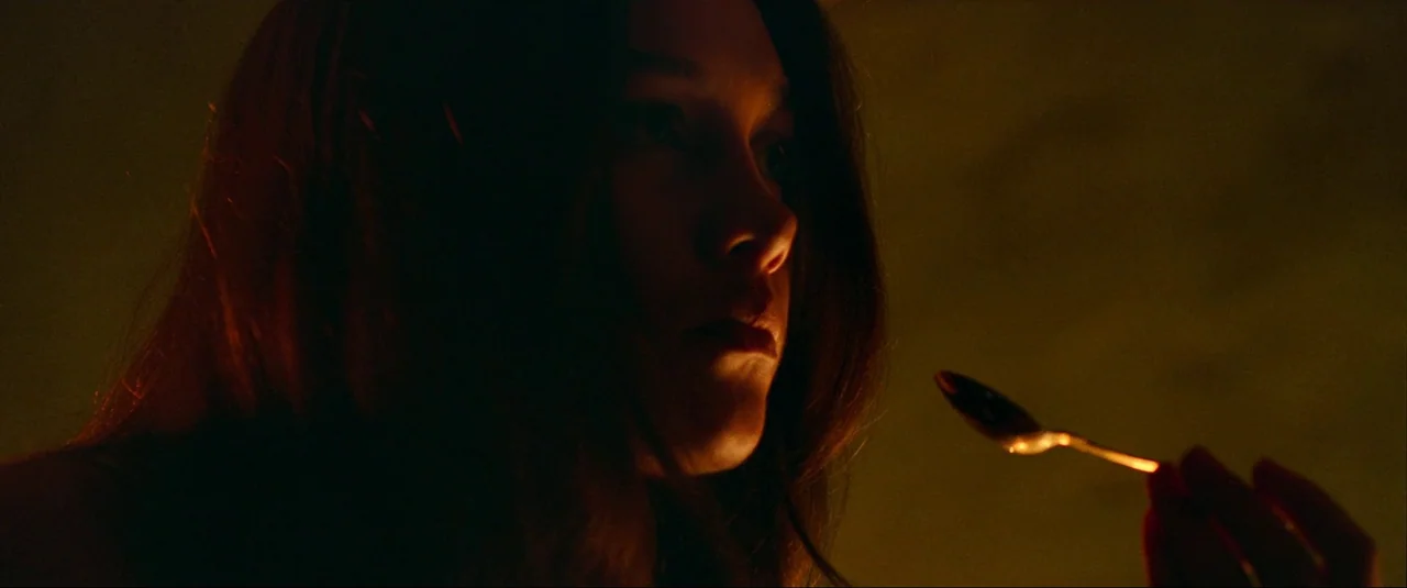 The Wonder (2022) movie still: closeup — The frame shows a dark interior space with warm ambient lighting. A woman holds a metallic spoon near her…