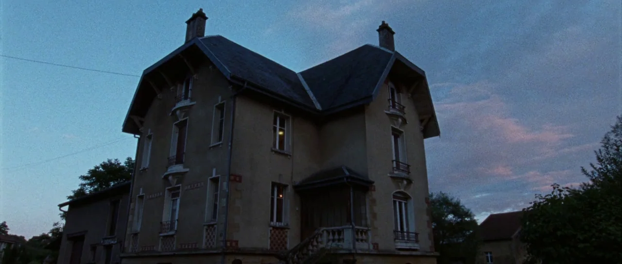 Alléluia (2014) movie still: establishing — A two-story residential building with stone detailing sits under a blue dusk sky. The building remains…