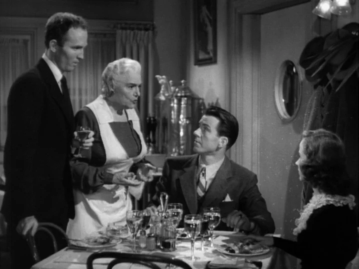 The Seventh Victim (1943) movie still: medium — A formal dining room setting features a table set with wine glasses and plates. Four adults engage…