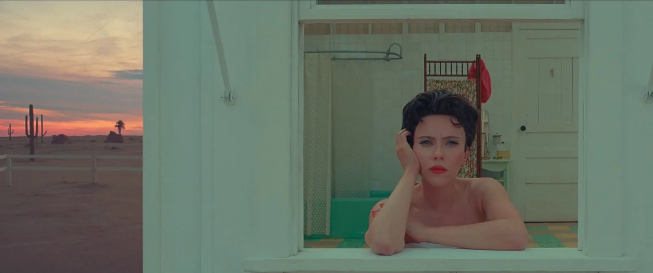 Asteroid City (2023) movie still: medium — A woman leans out of a wooden cabin window facing a flat desert landscape. Behind her is a bathroom…